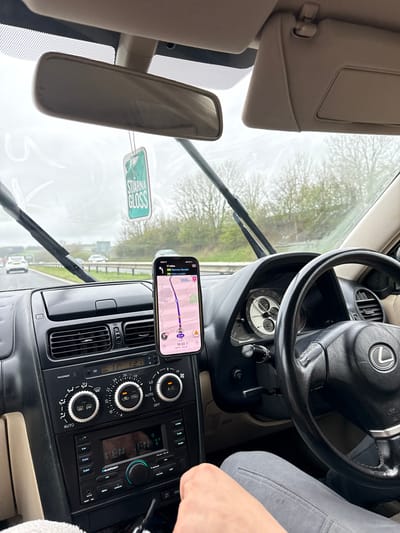 The image shows the interior of a Lexus car, likely a Lexus IS model, with the focus on the dashboard area. The car is equipped with a Blaupunkt PALMA 190 BT car stereo system and climate control dials, and the Lexus logo is visible on the steering wheel. The windshield wipers are active, suggesting it is raining.
A smartphone displaying a navigation app is mounted on the dashboard. The screen shows a route from an unknown location to Barrow, Kendal, and Kirkby Lonsdale.
A "Stjarna Gloss" car freshener hangs from the rearview mirror. The image was likely taken in the United Kingdom, based on the navigation information and the presence of road names. The navigation app reads "17 miles" to Barrow, Kendal, and Kirkby Lonsdale, and the time displayed is 15:33. The navigation app reads 1071.54 miles.
While the navigation app indicates the car is en route to Kendal and Barrow, and Kirkby Lonsdale, there is no visual indication of being in Lancaster, UK, visible in the image. A hand appears on the right side, likely belonging to the driver or a passenger.