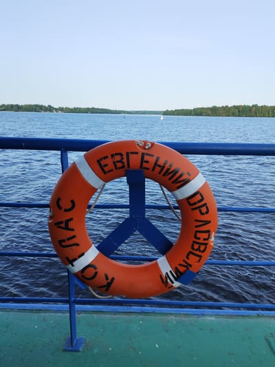The image shows an orange lifebuoy attached to a blue railing on a boat, with a calm blue body of water in the background. The lifebuoy has black Cyrillic text written on it, which reads "ЕВГЕНИЙ" on the top part and "СПАС" and "ОРАСВЕР" on the bottom part. The water is rippling gently, suggesting a peaceful day. In the distance, a tree-lined shore is visible under a clear, light blue sky, indicating fair weather and likely daytime. The boat's deck, partially visible in the foreground, is painted green. The scene evokes a sense of safety and tranquility associated with being on the water. The lifebuoy is a prominent object, highlighting the potential for water-related activities or the context of being on a vessel. The overall mood is serene, with the natural elements of water and sky dominating the backdrop. The text on the lifebuoy may indicate a name or a message related to rescue. The location context is Bessolovo, Russia.