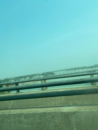 A view from a high vantage point, possibly a bridge or tall building. The sky is a light blue, fading towards the horizon. A metal truss structure runs along the left side, with parallel dark pipes and a concrete wall to the right. There's a narrow body of water between the truss structure and the pipes. The scene has a slightly hazy or muted quality.