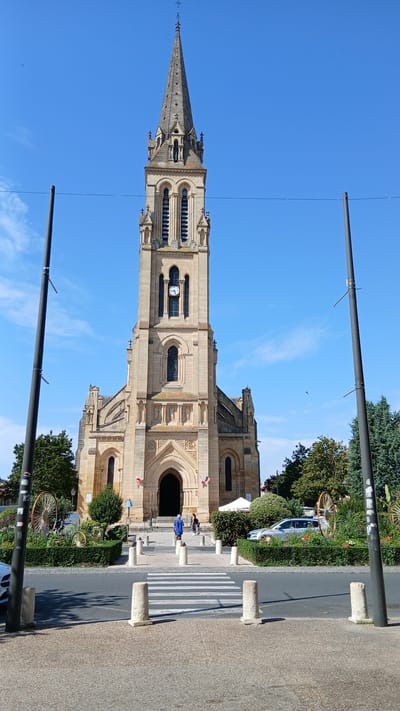 The image captures a front view of the Église Saint-Jacques in Bergerac, France. The church features a tall, ornate bell tower with a spire, a large arched entrance, and decorative stonework. The building is constructed from light-colored stone. In front of the church, there is a paved area with a pedestrian crossing, surrounded by low bollards. To the left and right of the church entrance, there are manicured hedges and decorative elements resembling large wagon wheels. A few people are visible near the entrance, and a silver car is parked behind the hedges on the right. Two tall, dark lampposts with wires running between them frame the scene. The sky is clear and blue with a few scattered clouds.