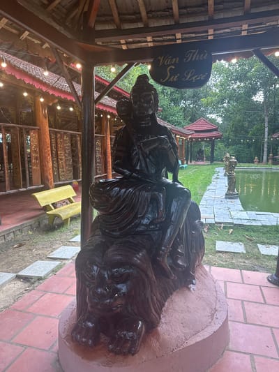 This media file depicts an outdoor scene in Suối Câu, Vietnam. The primary subject is a large, dark wooden sculpture of what appears to be a person, possibly in a reclining or seated position. The sculpture is intricate and detailed, with visible textures and flowing lines that suggest clothing and hair. It is placed on a circular, reddish-brown concrete base.
In the background, there's a tranquil pond with green water, lined with a stone pathway. Further back, a traditional Vietnamese gazebo with a red, multi-tiered roof is visible. Other decorative elements, including a statue of a person holding something above their head and string lights, are scattered throughout the scene, hinting at a festive or contemplative atmosphere. The overall setting is lush and green, with trees and grass surrounding the structures. The presence of string lights suggests it might be evening or a dimly lit area.
To the left of the sculpture, a dark, oval-shaped sign with gold lettering is hanging from a wooden structure. The visible text on the sign reads "Văn Thù Sư Lợi". The wooden structure appears to be part of an open-air building with a covered roof and decorative beams.
The image has a vertical orientation. The lighting is somewhat diffused, suggesting it may be overcast or late in the day. The scene evokes a sense of cultural richness and peacefulness, typical of a temple or a culturally significant garden.