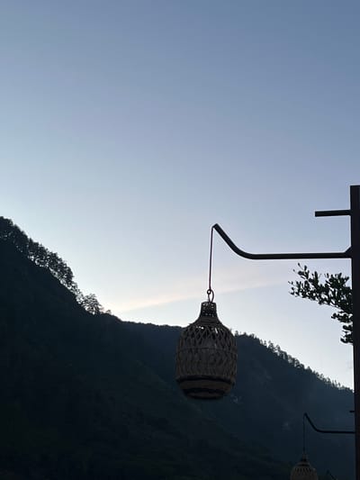 The image depicts a serene outdoor scene with a focus on a decorative woven lantern hanging from a metal pole. The lantern, made of natural fibers, is a prominent object in the foreground, with its intricate weave and a visible light bulb inside. Behind the lantern, a dark, tree-covered mountain slopes upwards against a clear, pale blue sky. The mountain silhouette suggests a natural landscape, possibly at dusk or dawn, given the soft lighting. The location is identified as Merodot, Indonesia. The scene is tranquil and devoid of human presence.