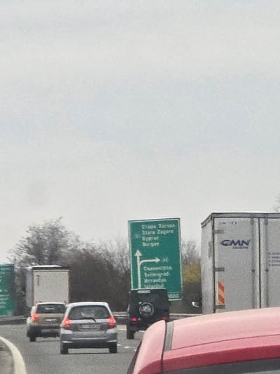 The image depicts an outdoor scene on a multi-lane motorway under an overcast, light grey sky. In the immediate foreground, the upper part of a red vehicle is visible, indicating the perspective of the observer from within this vehicle. In the midground, traffic is present in two lanes. The left lane contains a silver hatchback followed by a gold-colored hatchback, and a white semi-trailer truck further back. The right lane shows a black SUV, possibly a Mercedes G-Wagen, with a spare tire mounted on its rear. To the far right, a large white semi-trailer truck bearing "CMN" branding is visible. A prominent green highway sign stands on the right side of the road, displaying "Стара Загора" (Stara Zagora) and "Бургас" (Burgas) with a straight arrow, and "А1" (A1 motorway), "Свиленград" (Svilengrad), and "Истанбул" (Istanbul) with a right-pointing arrow, indicating an exit. A line of bare trees and shrubs forms a natural barrier along the left side of the motorway. No individuals are clearly discernible. The scene is located on the A1 Trakiya Motorway in Bulgaria, at the interchange providing directions to Stara Zagora, Burgas, Svilengrad, and Istanbul, geographically situated near the village of Cherna Gora in Stara Zagora Province, Bulgaria.