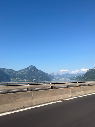 The image presents a static, clear, outdoor scene, likely captured from a highway bridge or elevated road in Beckenried, Switzerland. In the immediate foreground, a dark asphalt road surface with a distinct white lane marking on the right leads to a light grey concrete barrier. Beyond this barrier, a dark metal safety railing is visible, followed by the calm, deep blue waters of Lake Lucerne (Vierwaldstättersee) extending horizontally. The mid-ground features a large, conical, forested mountain, consistent with the Buochserhorn, covered predominantly in dark green trees with some lighter green patches. The town of Beckenried is visible at the base of this mountain, along the lake's shoreline, characterized by numerous white and light-colored buildings. In the background, additional rugged, treed, and rocky mountains recede into the distance, with faint white cloud formations near their peaks on the right. The upper half of the frame is dominated by a clear, bright blue sky. No individuals, animals, or moving objects are present in the scene.