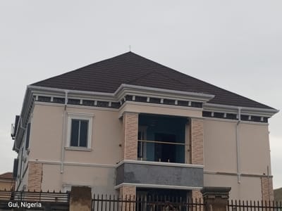The image shows a two-story building under construction or recently completed in Kuje, Nigeria. The building has a dark brown tiled roof with white trim and gutters. The exterior walls are painted a light cream color, with some sections featuring a brick-like texture in a lighter shade. There is a prominent balcony on the upper floor, with tiled pillars and a blue interior. The balcony is enclosed by a railing. Below the balcony, there is a gray concrete section. On the left side of the building, there is a large, dark-framed window. A metal fence with vertical bars is visible in the foreground, partially obscuring the lower part of the building and a concrete pillar. The sky is overcast and gray. The bottom left corner of the image has a watermark that reads "Gui, Nigeria". There are no people visible in the image.