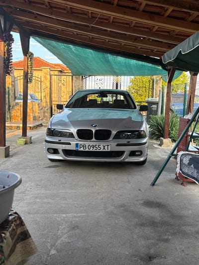 This image captures a silver BMW E39 5-series sedan parked head-on under a wooden carport structure in Vinitsa, Bulgaria, likely during late afternoon or early morning hours, indicated by warm, directional sunlight.
The primary subject is the silver BMW E39, featuring aftermarket black-housing headlights and an M-Sport style front bumper. A script, possibly "M-Performance," is faintly visible on the hood above the driver's side headlight. The vehicle displays a Bulgarian license plate, "PB 0955 XT," positioned centrally on the front bumper. The car is situated on a concrete driveway, which shows minor cracks and dark stains.
The carport overhead consists of natural wood beams and rafters, covered by a green mesh shade net. On the left foreground, artificial purple and white floral decorations hang from a wooden pillar. A white plastic basin, partially visible, rests on a patterned cloth to the lower left. In the right foreground, a dark green metal frame (possibly from a swing or chair) extends into the frame. An off-white sneaker is visible on a brown woven chair or stool, draped with a dark patterned fabric.
The background on the left reveals a building with an orange-tiled roof and walls clad with multi-colored, irregularly shaped stone. A decorative, ornate metal gate with gold or brass-colored scrollwork stands in front of this building. A light-colored vehicle is partially visible behind this gate.
The background on the right features lush green foliage and trees behind a dark metal fence, followed by a green mesh fence. A dark gray waste bin, labeled "AREA Recycling," is visible next to a plant with spiky leaves. Hints of a blue structure are discernible further in the distance on the right.
No individuals are visible within the frame.