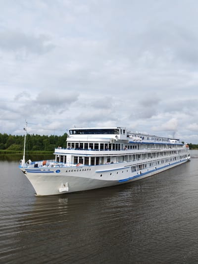 A large, white river cruise ship named "АЛЕКСАНДРА" is depicted in the foreground, sailing on a calm body of water. The ship has multiple decks with numerous windows and is adorned with blue trim along its sides. Several people are visible on the upper decks, observing the surroundings. The ship is moving from left to right in the frame. In the background, a lush green forest lines the opposite bank of the river. The sky is overcast with dramatic, grey clouds. The scene is set in the city of Kargulino, Russia.