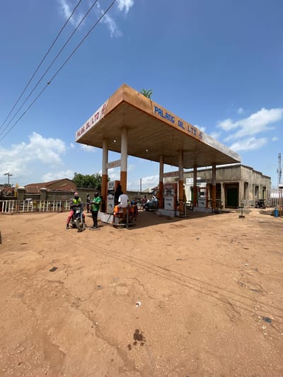 This is a wide-angle, eye-level shot of the Palang Oil Ltd. gas station in Tangu, Nigeria. The gas station has a large, weathered orange and white canopy supported by concrete pillars. Several gas pumps are visible under the canopy, with one being used to fuel a motorcycle.

In the foreground, the ground is unpaved, dusty, and reddish-brown, with some oil stains. In the midground, a man on a motorcycle, wearing a bright yellow vest, is interacting with a man in a green shirt. Another person in a white shirt is seated on the side of the gas pump. A few other people are visible in the background, near the pumps and a small building to the right of the gas station. The building is made of concrete and appears to be part of the gas station's facilities. A white metal fence runs along the left side of the property, behind which are some residential or commercial structures. Overhead, power lines stretch across the frame, and a bright blue sky with scattered white clouds dominates the upper portion of the image. The overall scene depicts a functional but somewhat rustic gas station in a Nigerian town.
