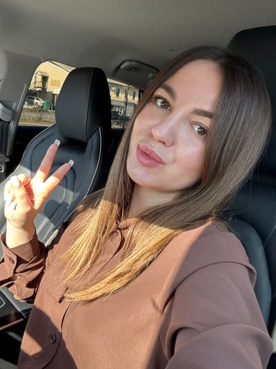 A female individual is depicted seated in the driver's seat of a vehicle in Likino-Dulyovo, Russia, during daylight hours on a clear, sunny day. The woman has long, straight brown hair and light skin, with dark brown eyes and full lips. She is wearing a brown, long-sleeved collared shirt with visible buttons and a tie detail at the neckline. Her left hand is raised, forming a "peace" gesture with her index and middle fingers extended; her fingernails are manicured with light-colored tips. She is looking directly towards the camera. Direct sunlight illuminates the right side of her face and hair, casting prominent shadows across her chest and left arm. The interior of the vehicle features a black leather passenger seat directly behind her. Through the driver's side window, an urban street scene is visible, comprising multi-story buildings, one featuring an external staircase, and a light-colored vehicle on the roadway.
