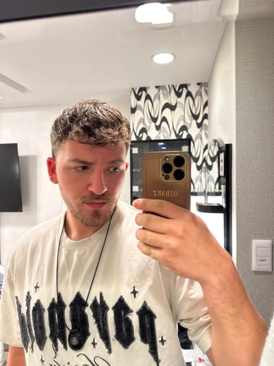 The image shows a man standing in front of a mirror. He is wearing a white t-shirt with a black pendant necklace and a black graphic design. His right arm is extended in front of him, holding a gold iPhone with a black case that says "OSBWRZ" in reverse. The man has short brown hair and a light beard. He is looking directly at the camera. The location is a bathroom in a hotel, located at 35.675 latitude and 139.741 longitude in Chiyoda City, Japan. The background shows a white wall with a black and white abstract design and a shelf with various toiletries. There are two white ceiling lights above the man. The man appears to be taking a selfie.