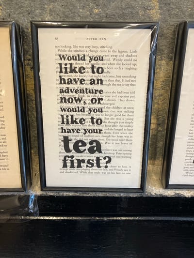 A framed print, approximately 10x16 inches, is displayed centrally on a dark, flat surface in Ambleside, United Kingdom. The print features a page from a book, identified by the visible header "PETER PA" and page number "88", which is "Peter Pan". Specific words within the text are enlarged and bolded to form the question: "would you like to have an adventure now or would you like to have your tea first?". The print is enclosed in a narrow, glossy black frame and protected by transparent, crinkled plastic shrink-wrap, which shows reflections from ambient lighting. Partially visible identical framed prints are positioned to the left and right, indicating a series of similar items. The background behind the frames is a textured, mottled dark beige/grey surface. No people or active interactions are present in the scene.