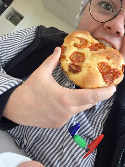 A person with glasses is taking a bite out of a round bread topped with tomatoes. The person is wearing a grey and white striped shirt and a black vest. In the shirt pocket are a red pen, a green object, and a blue object. The location cannot be determined as being in Mont-Saint-Martin, France based solely on the visual information.