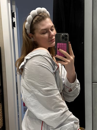 The scene is located indoors in the city of Bakcha-Sarai, Russia. An adult female is depicted from the upper torso upwards, taking a mirror selfie with a smartphone held in her right hand. She has light brown hair styled down and wears a light grey, segmented headband. Her left ear displays a silver-toned, multi-clustered earring. She is dressed in a light-colored, striped pajama-style top with red piping, which is pulled off her left shoulder. Her eyes are closed or looking downwards, and her head is slightly tilted. The smartphone is dark-colored, featuring a triple-lens camera array and a pink pop-it style accessory adhered to its back. A white cable extends from the phone. The background visible in the mirror includes a white door frame with dark hinges on the left, a dark surface behind the subject, and a light grey tiled or paneled wall section on the right. A faint reflection of a woven basket is discernible in the lower-left corner near the door frame.