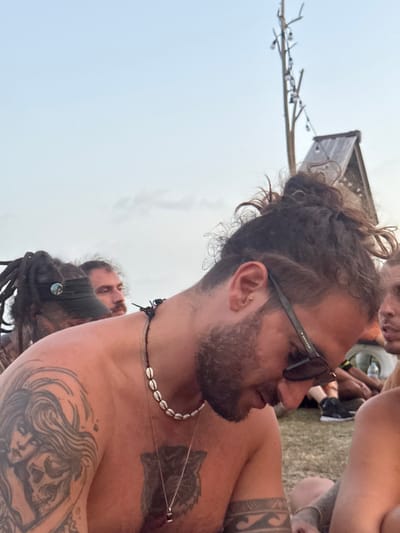An outdoor scene in Ban Pang Pring, Thailand, depicts a group of individuals in what appears to be a casual gathering. The foreground is dominated by a shirtless male with curly brown hair styled in a top knot, a beard, and dark sunglasses. He wears a necklace of white seashells and a silver chain with a small pendant. Visible tattoos include a detailed rendering of a woman embracing a skull on his left shoulder and arm, and a dark, possibly animalistic, figure on his chest. His head is tilted downward.
To his left and slightly behind him, a second male is partially visible, wearing dreadlocks and a dark visor with a circular badge. Behind these two, a third male with a mustache is visible, looking toward the right. Additional indistinct figures are present in the middle ground and background, some appearing to be seated on the ground, which is covered in grass or dirt.
The background features a light blue-grey sky, suggesting late daylight conditions. In the upper right, a rustic wooden structure with a sloped roof is present, alongside a tall, bare pole or tree trunk adorned with string lights.