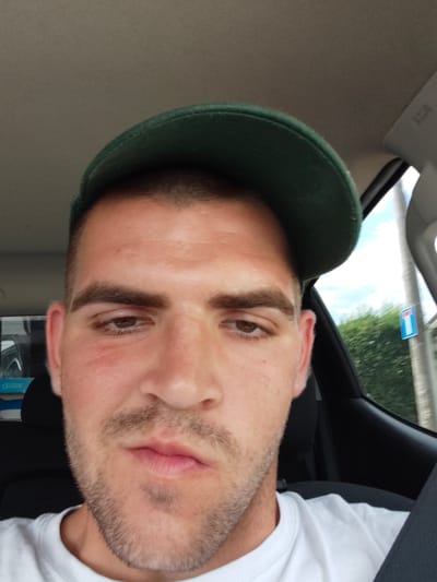 The image is a selfie of a man inside a car, likely in Lombardore, Italy. The man is wearing a green baseball cap and a white t-shirt. He has short, dark hair, brown eyes, and a light stubble on his face. His lips are slightly parted. The background shows the interior of a car, with a glimpse of the outdoors through the window, suggesting it's daytime. Trees and a road sign are visible outside, indicating a rural or suburban setting. The man's expression is neutral. There is no visible text in the image.