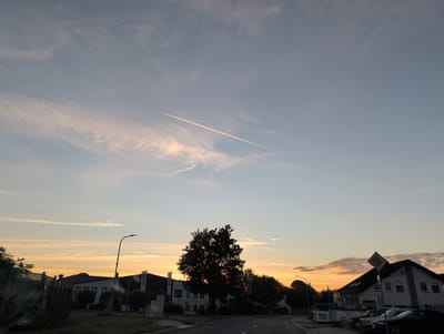 The image depicts a street scene in Polch, Germany, during sunset. A thin contrail from an airplane stretches across the pale blue sky, which is tinged with orange and pink hues from the setting sun. Scattered clouds, some wispy and others more substantial, are visible. In the foreground, a dark silhouette of a large tree stands out against the colorful sky. To the left, a series of industrial-style buildings with dark roofs and white facades are visible, one with a prominent street lamp in front. To the right, a multi-story building with a dark roof and white walls is partially visible, with several cars parked in front. The street is paved and appears to be a road, with white lane markings. The overall impression is a peaceful evening scene in a semi-urban or industrial area.