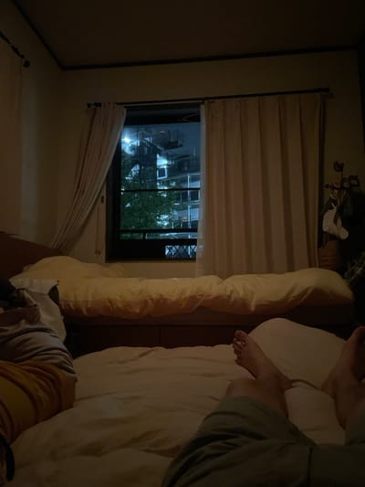 A person's legs are visible in the foreground of the image, lying on a bed in a dimly lit room in a residence located in Katsushika City, Japan. The person is wearing khaki shorts and has bare feet. The bed is made up with a white comforter and two pillows. A second bed with a yellow comforter and two pillows is visible in the background. The room has a window to the left of the image that is open and has white curtains drawn slightly back. The window looks out onto a tree and an apartment building. The room has dark brown trim and a white ceiling. The image is taken from a low angle, looking up at the person's legs.