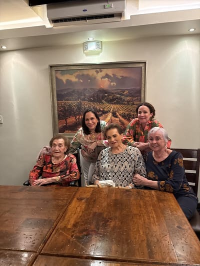 A group of five women of varying ages are posing for a photo behind a wooden table. The wall behind them is painted a light color and features a landscape painting in a gold frame. Above the painting, near the ceiling, is an air conditioning unit and a light fixture. The women are dressed in patterned tops, and they are all smiling. It is unclear from the image whether the scene is in Tijuana, Mexico, but the description includes the elements visible in the picture.