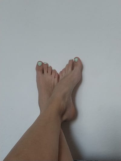 This is a close-up, waist-up photograph of a person's bare feet crossed against a plain, light-colored wall, likely indoors. The feet are positioned in the lower half of the frame, with the soles facing slightly upwards. Both feet are neatly presented, with light green nail polish applied to the toenails, some adorned with small, clear rhinestones. The lighting is soft and diffused, creating subtle shadows that highlight the contours of the feet and legs. There is no visible interaction or action; the focus is solely on the aesthetic presentation of the feet. The background is a simple, textured wall with no discernible features, suggesting a domestic or studio setting. There is no information in the image to determine the location in Dobev, Czechia.