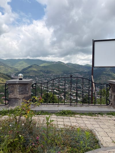 In the city of Goris, Armenia, a scene unfolds with a panoramic view of a sprawling town nestled within a valley, its buildings characterized by red-tiled roofs. The town is surrounded by rolling, verdant mountains under a sky of dramatic, heavy clouds. In the foreground, a stone-built pillar with an ornate light fixture stands guard, its rough texture contrasting with the smooth, patterned paving stones that extend towards the viewer. A black wrought-iron fence, with intricate scrollwork, separates the elevated viewpoint from the town below, suggesting a park or a private residence. Lush vegetation, including small bushes and flowering plants with pink and white blooms, adds touches of color to the immediate foreground. To the right, a blank white rectangular sign is prominently displayed on a metal pole, its purpose undefined. The overall impression is one of a picturesque landscape overlooking a historic Armenian town.
