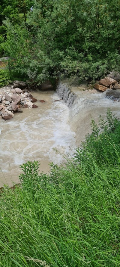 The image shows a natural scene with a small waterfall. The water is cloudy, suggesting it may be from recent rain or a swollen stream. Rocks and pebbles line the stream bed, and lush green grass and trees surround the area. The environment appears to be a natural landscape, possibly a park or a rural area. There are no people or man-made structures clearly visible in this portion of the image. The scene is tranquil and depicts a natural water feature.