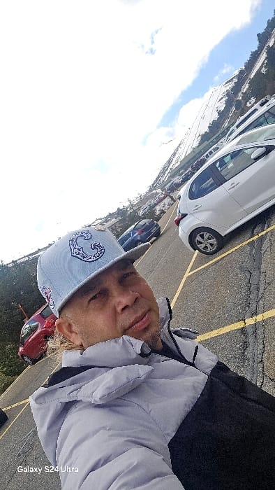 An adult male is depicted in a close-up selfie, positioned at the lower left of the frame. He wears a light grey baseball cap featuring a white, ornate, stylized design resembling a letter 'C' on the front. His face shows light stubble, and his expression is neutral. He is dressed in a two-tone puffer jacket, light grey on the upper half and dark grey or black on the lower half. The image is taken outdoors in what appears to be a parking lot located in a mountainous region of Rascafría, Spain.
The background shows an asphalt surface with yellow painted parking lines and visible cracks, sloping upwards from left to right. Several vehicles are present: a white hatchback car is parked immediately behind the man, partially visible; a blue car is further up the slope; and a red car is partially visible on the far left. Beyond the parking area, a steep, snow-covered mountain slope is visible, interspersed with dark green evergreen trees. The sky is partially cloudy with patches of blue. The bottom-left corner of the image contains a "Galaxy S24 Ultra" watermark. The entire scene is brightly lit by natural daylight.
