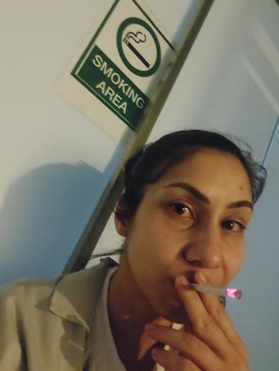 A medium-close, tilted indoor shot depicts a woman smoking a cigarette in a designated smoking area. The woman, of apparent Middle Eastern descent, holds a lit cigarette in her right hand near her mouth. Her dark hair is pulled back. She has brown eyes and fair skin. She is wearing a light grey top.
To the right of the woman is a rectangular sign with the words "SMOKING AREA" in green lettering. Below the text is a green and white graphic depicting a cigarette inside a circle with a slash through it, with smoke emanating. The background is a light, cool-toned wall. The scene appears to be indoors, possibly in a designated smoking space in a public facility or restaurant. The city location of Altdorf, Germany is not identifiable from the visual elements.