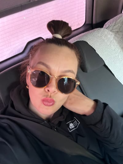 One female subject is visible from the chest up, positioned inside a vehicle in Gol'yany, Russia, taking a self-portrait. She has brown hair styled in a high bun with a light elastic. The subject wears light-framed sunglasses with dark lenses and a dark hooded garment displaying a white Kappa logo on the left chest. Her lips are puckered, and a small gold-colored hoop earring is visible on her left ear. Her left hand rests against the side of her face, and a dark seatbelt crosses her right shoulder. The background shows a car window with a pinkish tint and a blurred white knitted blanket draped over an adjacent seat.