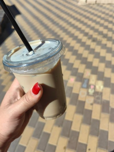 An outdoor, eye-level shot captures a person's hand holding a clear plastic cup filled with a light brown beverage and ice. A black straw extends from the cup's lid. The hand, with a bright red nail polish on one fingernail, is positioned in the lower-left quadrant of the frame. The background consists of a paved ground with a repeating pattern of beige and grey rectangular tiles, illuminated by diagonal shadows. Some faint chalk drawings, including a pink bunny and other indistinct shapes, are visible on the tiles in the middle ground. The scene appears to be in a public outdoor space within the city of Risovyi, Russia.