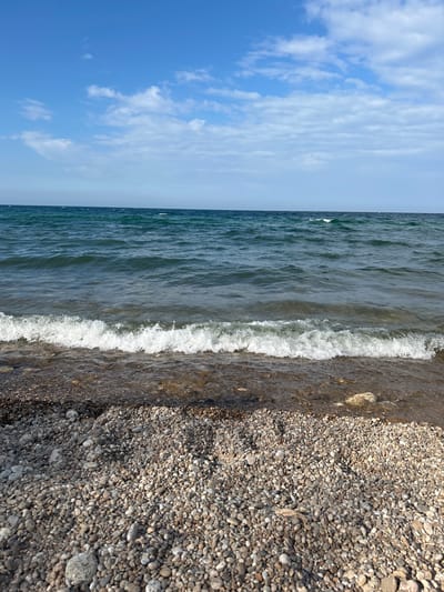 The image depicts a clear day at a pebble beach on the shore of Lake Baikal, near the village of Tolbazikha, Russia. The sky is a vibrant blue, with scattered white clouds. The water of Lake Baikal is a deep, dark teal color, with small waves lapping at the shore. The waves are breaking, creating white foam as they reach the pebbles. The beach itself is covered in smooth, grey and brown pebbles of various sizes. The overall impression is one of natural beauty and tranquility.