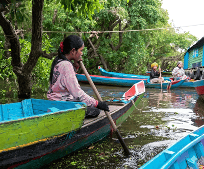 In a narrow waterway in the city of Soutr Nikom, Cambodia, several colorful boats are navigating through lush green foliage. In the foreground, a young woman with dark hair tied back with a red ribbon is seated in a brightly painted blue and green wooden boat. She is wearing a pink sweatshirt with a graphic design and dark gloves, and she is actively rowing with a wooden oar. The water is dark and reflective, with scattered green plant life. Behind her, two other women are also in boats. One woman, wearing a yellow headscarf, is rowing a blue and red boat, while another woman, in a patterned top and headscarf, sits in a similar boat behind her. To the right, part of a blue and green stilt house is visible, suggesting a stilted village setting. The environment is dense with trees and greenery, creating a shaded and natural atmosphere.