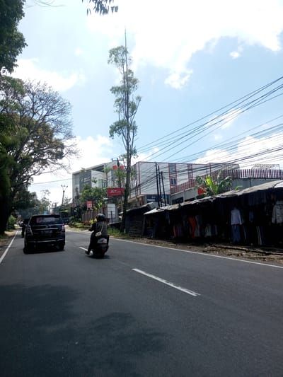 The image shows a street in Kioumaknao 2, Laos, featuring a black car and a motorcycle traveling on a paved road with a white line dividing the lanes. Buildings are visible on the right side of the road, including one with "J&T Express" signage. There is a covered area along the roadside that appears to be a market or stall selling clothing. Overhead electrical wires crisscross the sky. Trees line the left side of the road. The sky is blue with scattered clouds.