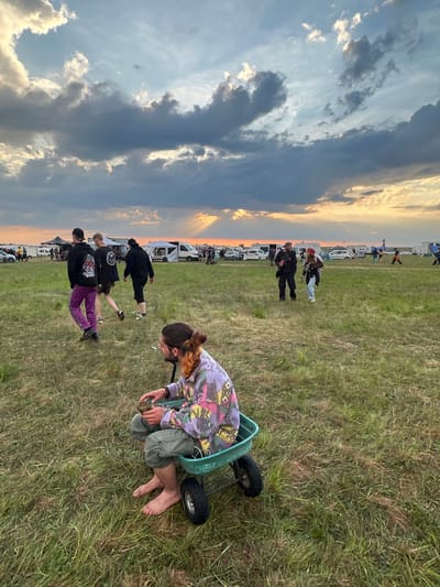 The image, taken outdoors in Bengy-sur-Craon, France, shows a grassy field setting at dusk. A young individual, seen from the side, is kneeling inside a small, wheeled, green cart. They are wearing a tie-dye top, green pants, and are barefoot. Their hair is pulled back. They appear to be examining or adjusting something in front of them. In the background, multiple individuals stand and walk in the same direction as the cart, with a line of parked vehicles visible further back. The sky in the background features a sunset with hues of orange and yellow. The lighting appears natural.