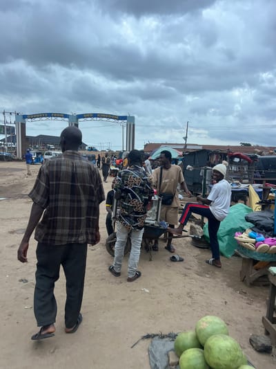 The image depicts a street scene in Oko Amakom, Nigeria, under a cloudy sky. In the foreground, a man in a brown plaid shirt and dark pants walks away from the camera. To his right, several other individuals are engaged in various activities. One man in a tan shirt and shorts stands near a cart, while another man in a white t-shirt and dark pants with red stripes playfully poses, with his leg raised. In the background, an archway with the inscription "GOODBYE FROM OKO KINGDOM" is visible, marking an entrance or exit. The environment appears to be a market area, with stalls, goods displayed on tables, and vehicles, including a tricycle, in the distance. The ground is sandy and dusty. A pile of green gourds is situated in the lower right corner of the frame. The overall atmosphere suggests a bustling, everyday scene in a Nigerian town.