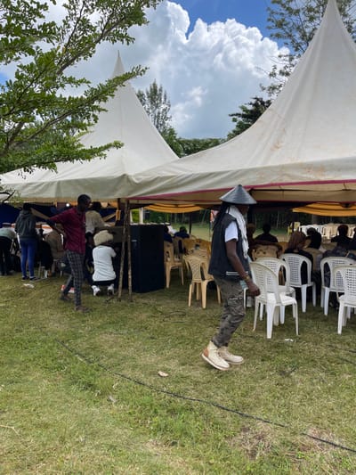 The image captures an outdoor event taking place on a bright, sunny day, likely in Makuyu, Kenya. The scene is set on a vibrant green lawn, under two white canvas tents providing shade. Large, fluffy white clouds are visible against a clear blue sky, with lush green trees and foliage framing the background and foreground, indicating a natural, open-air setting.
Several people are gathered at the event. A prominent male figure stands in the foreground, wearing a distinctive conical hat, a dark vest over a white t-shirt, camouflage-patterned pants, and light-colored boots. He is positioned near the opening of one of the tents. Within and under the tents, numerous white plastic chairs are arranged, with several people seated, some partially visible or facing away from the camera. One person in a maroon shirt is seated closer to the foreground.
The setup, including the tents, chairs, and large black speakers visible under one of the tents, suggests an organized outdoor gathering such as a social function, meeting, or celebration. Wires are present on the grass, likely connecting to the sound equipment. The overall atmosphere appears relaxed. The bright daylight and clear skies indicate good weather conditions for the outdoor occasion. There is no discernible text visible in the image.