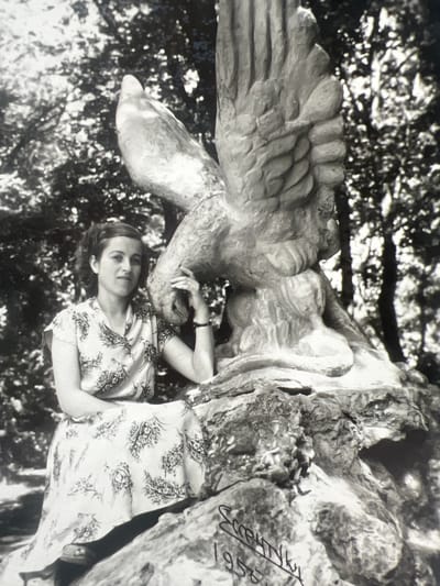 A black and white medium-shot photograph shows a woman sitting on a rock next to a large eagle sculpture. The woman is wearing a light-colored floral print dress and has dark hair styled in soft waves. She is looking towards the viewer and resting her right hand on the eagle's beak. The eagle sculpture is detailed, with visible feathers and talons. The background consists of trees and foliage, suggesting an outdoor setting. In the lower right corner of the image, handwritten text is visible, appearing to be a name and the year "1958". The location is identified as Дружба Снт, Russia.