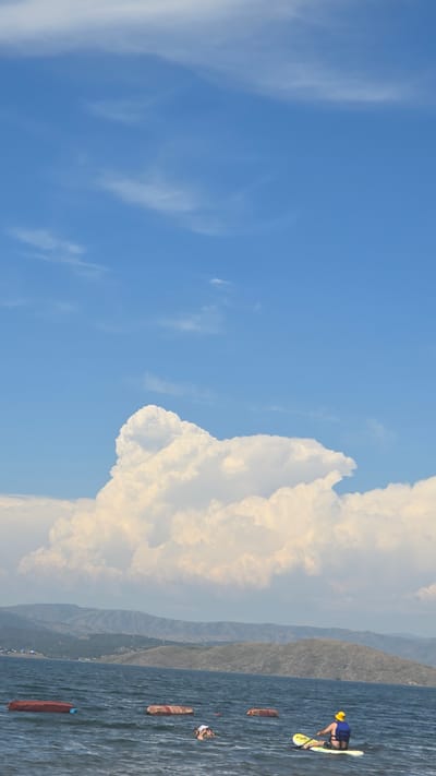 The image depicts a serene lake scene with a person on a paddleboard in the foreground. The person is wearing a blue life vest and a yellow hat, and is facing away from the camera, actively paddling. Several red floating objects are scattered in the water. In the background, a vast body of water stretches towards a distant, hilly landscape under a bright blue sky filled with large, fluffy white clouds. The scene appears to be set in Manat, Kazakhstan, a location known for its natural beauty and recreational water activities.