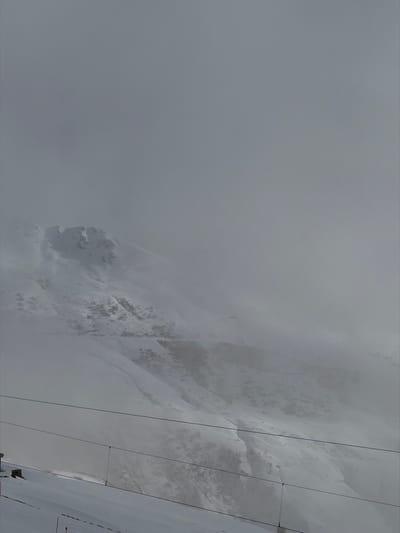 The image displays a snow-covered mountain landscape in Andermatt, Switzerland, under heavy cloud cover and fog. The upper two-thirds of the frame are dominated by a uniform, light gray sky obscured by dense fog or low-hanging clouds, significantly reducing visibility. Below this, snow-dusted mountain slopes descend, revealing rugged terrain with exposed rock formations and patches of bare earth. A faint, winding path or road is discernible traversing a distant slope through the mist. In the immediate foreground, two dark, parallel cables, consistent with ski lift infrastructure, stretch horizontally across the lower portion of the image. A small, snow-covered structure, possibly a lift tower base, is visible near the lower-left corner, with a few red and white marker poles partially visible in the snow below the cables. No individuals or active movements are present.