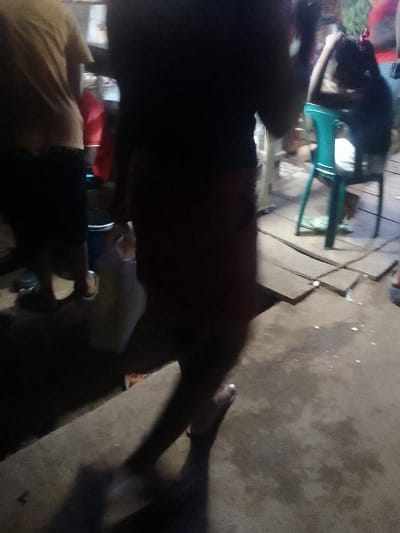 A blurred scene shows several people and a green plastic chair. One person, partially in frame, wears a tan shirt and dark shorts. Another figure is mostly obscured, wearing a dark top and reddish-brown skirt. A small child with a red bow sits in the chair. The foreground features a concrete surface with long shadows, suggesting an evening or nighttime setting. A plastic bag sits near the first person's feet.