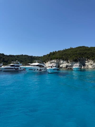 This is a photograph taken from a body of water, likely the sea, looking towards a coastline. The water in the foreground is a vibrant, clear turquoise blue with gentle ripples. In the midground, several white motor yachts are moored. The largest yachts are on the left, with sleek, modern designs. Further to the right, there are smaller yachts and a rocky coastline with lush green vegetation covering the slopes above. The sky is clear and a bright blue, indicating a sunny day. The overall scene depicts a beautiful coastal landscape with luxury boats in a serene, tropical-like setting, consistent with locations like Antipaxos, Greece, known for its stunning turquoise waters and secluded coves.