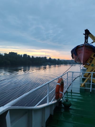The image depicts a serene scene from the deck of a ship on a river, likely during dusk or dawn, with a clear view of the town of Ivanov Bor, Russia, along the riverbank.
In the foreground, the green deck of the ship is visible, glistening with moisture, suggesting recent rain or the morning dew. A white railing runs along the edge of the deck. A bright orange life preserver ring hangs on the railing. To the right, a yellow crane structure is present, supporting a red lifeboat covered with a blue tarp. The lifeboat has some Cyrillic text visible on its side, which appears to be part of its identification. A green valve or cap is also visible on the deck.
The river stretches out ahead, with gentle ripples on its surface reflecting the soft, warm hues of the sunset or sunrise. The water appears calm, indicating a peaceful journey. On the far bank, a dense line of dark green trees forms the natural landscape bordering the river. Beyond the trees, the town of Ivanov Bor is visible with scattered houses and buildings. A telecommunication tower or antenna is discernible on the horizon to the right.
The sky above is a gradient of soft blues and grays, with wispy clouds that catch the light from the setting or rising sun, creating a warm band of orange and yellow near the horizon. The overall atmosphere is tranquil and somewhat melancholic, evoking a sense of quietude and reflection. There are no people visible in the image.