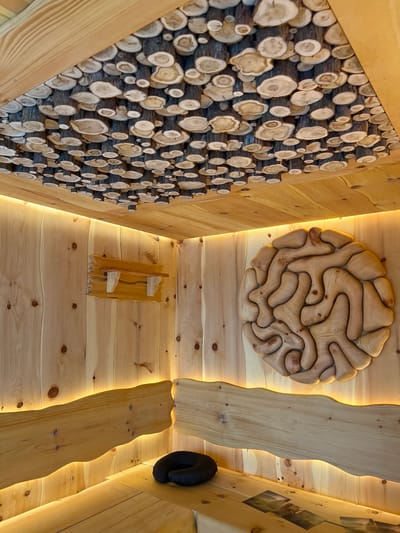 The image depicts the interior of a room constructed entirely of wood, likely a sauna or spa facility in Yakunino, Russia. The ceiling is adorned with a mosaic of circular wood slices of varying sizes, creating a natural, organic pattern. The walls are paneled with vertical wooden planks, and accent lighting runs along the top edges of the walls, casting a warm, yellow glow.
Dominating the right wall is a large, circular wooden sculpture with an intricate, maze-like design. On the left wall, a small, rustic wooden shelf is mounted. Built-in wooden benches line the lower portion of the room. On one of the benches, a black neck pillow rests, and several pieces of paper, possibly brochures or menus, are laid out. The overall aesthetic is rustic and natural, emphasizing the use of wood in both construction and decoration. There are no people visible in the image.