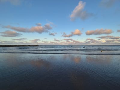 The image shows a beach at sunset, likely located in Boca del Río, Mexico, given the landscape and coastal setting. The sky is predominantly blue with scattered white and pink-tinged clouds. The sea is calm, reflecting the colors of the sky and clouds. Several people are visible in the water, some appearing to be swimming. A yellow marker or buoy is floating on the water's surface. In the distance, a stone pier or jetty stretches into the sea, with a lone individual standing at the end. The foreground consists of the wet sand of the beach, reflecting the sky and sea. The overall scene conveys a serene and peaceful atmosphere.