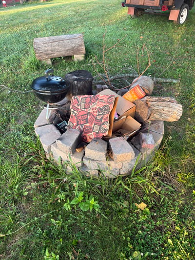This is a close-up, ground-level shot of a fire pit constructed from concrete blocks in a grassy yard, possibly in Holt, United States. The fire pit is filled with various items, including pieces of firewood, branches, and several cardboard boxes, one of which is a Pizza Hut box. A black charcoal grill sits on the edge of the fire pit, and a thick wooden log is positioned behind it. In the background, a small trailer with wooden planks is visible. The area is covered in green grass, with some patches of clover and weeds, suggesting an outdoor setting. The lighting indicates it is either early morning or late afternoon. No people or animals are present in the image.