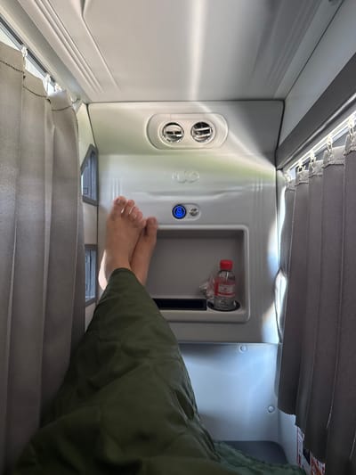 The image depicts the interior of a small sleeping compartment, possibly on a train or bus. The perspective is from someone lying down, with their bare feet and legs covered by a green blanket visible in the foreground. The compartment walls are white and appear to be made of plastic or a similar material. To the right, there are gray curtains that can be drawn for privacy. Above the person's feet, on the wall, are two circular air vents and a small niche containing a water bottle with a red cap. A blue illuminated button is also visible on the wall. The lighting suggests an artificial light source within the compartment, casting shadows and highlighting the textures of the materials. The overall impression is one of a compact, utilitarian personal space for rest. The location in Sông Lũy, Vietnam is not discernible from the image itself.