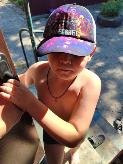 A young boy, shirtless and wearing black swim trunks, stands outdoors, holding onto a brown structure, possibly part of a playground. He wears a cap with a vibrant design featuring characters from the video game Minecraft and a galaxy-like pattern on the brim. A thin black necklace with a small gold cross is visible around his neck. The lighting suggests it is daytime, with strong sunlight casting shadows, indicating a sunny day. The background shows a grey gravelly surface and some greenery in a pot, hinting at a yard or outdoor recreational area. The boy's expression is somewhat solemn, and his eyes are mostly obscured by the cap's brim. Visible text on the cap reads "MINECRAFT". The scene appears to be located in Novolativka, Ukraine.