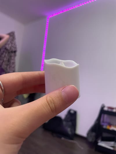 In a room illuminated by purple LED strip lights, a person's hand, adorned with a silver ring on the index finger, holds a small, white, artificial candle. The candle has a somewhat irregular, wavy top edge. The background is blurred, showing a wall with a patterned cloth hanging on it to the left, and dark furniture with shelves to the right. The location is identified as Borisova Griva, Russia.