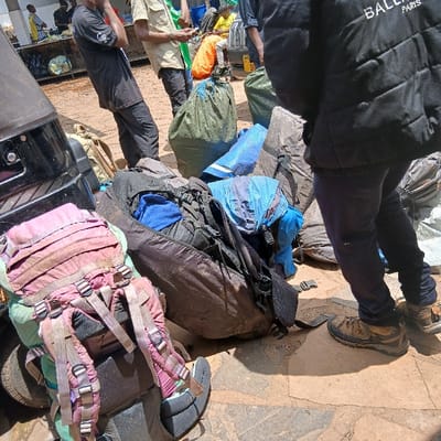 An outdoor scene in Mweka, Tanzania, depicts multiple individuals and a substantial collection of luggage on an uneven ground surface comprised of reddish-brown dirt and irregular stone pavers. In the immediate foreground, left, a large, dusty pink hiking backpack with multiple straps and buckles rests beside the rear quarter panel and tire of a dark-colored vehicle. Numerous other large bags, including dark duffel bags, green canvas-like sacks tied at the top, and blue tarpaulin-wrapped items, are scattered across the ground, forming a central pile. To the right, a person's lower torso and legs are visible, wearing a black puffer jacket with "BALENC PARIS" visible on the back, dark trousers, and brown hiking boots, standing with their right foot forward. In the middle ground, left-center, a person in a black t-shirt and black pants stands observing the scene. Further back, another individual in a light-colored shirt and dark pants stands, looking downward. Other partially visible individuals and a counter-like structure are present in the far background. The scene is brightly illuminated, indicating daylight.
