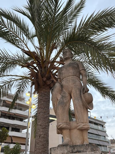 The image shows a stone statue of a man in Port d'Alcúdia, Spain, partially obscured by a palm tree. The statue, made of weathered stone, depicts a male figure in work clothes, holding a hat in his right hand and what appears to be a fishing net or similar item over his left shoulder. He stands on a stone pedestal. Behind the statue, a large palm tree with fronds extends into the frame. In the background, there are multi-story buildings with balconies and awnings, characteristic of a Mediterranean town. A lamppost with several yellow lights is visible to the left of the palm tree. Bunting is strung across the top of one of the buildings in the background. The sky is overcast.