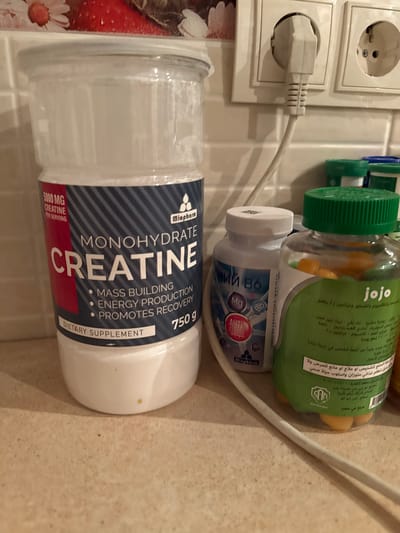 A static close-up image displays an arrangement of three product containers on a light-colored speckled countertop against a tiled wall in Ciorescu, Moldova. The primary object, positioned prominently on the left, is a tall, transparent plastic tub of "Miopharm CREATINE MONOHYDRATE" dietary supplement, containing white powdered substance. Its label specifies "5000 MG CREATINE PER SERVING," promotes "MASS BUILDING, ENERGY PRODUCTION, PROMOTES RECOVERY," and indicates a "750 g" quantity.
To the right of the creatine tub, a smaller white plastic bottle with a white screw-top lid is visible, featuring a label with the brand "Miopharm" and Cyrillic text, including "МГ В6." Adjacent to this is a transparent plastic bottle with a bright green cap, filled with numerous small, spherical, orange items, and labeled "jojo" along with Arabic text.
In the background, a light-colored tiled wall is equipped with a dual electrical outlet, from which a light-colored power cord extends downwards, partially obscuring the middle and right bottles. The upper left background shows a blurred section of what appears to be a decorative tile or wallpaper pattern featuring red strawberries and white daisies. The countertop surface exhibits minor variations in texture and a few small, yellowish flecks near the base of the creatine tub.